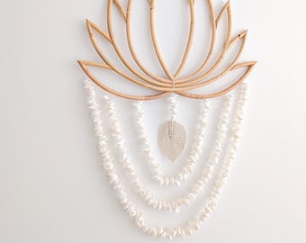 Puede incluir: Adorno de pared con forma de flor de loto, hecho de ratán tejido de color natural. Debajo del loto, cuelgan hilos de conchas blancas, con un acento central en forma de hoja texturizada. Una pieza decorativa de estilo bohemio.