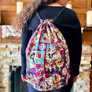 May include: A drawstring bag with a Wonder Woman comic book print. The bag is made of fabric and has a black drawstring. The bag is being worn as a backpack.