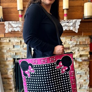 May include: A black and white polka dot tote bag with pink trim and a Minnie Mouse design. The bag has a pink bow on each side and a black Minnie Mouse head with a pink bow on each corner.
