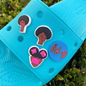 May include: Blue and white shoe charms with various designs, including a raised fist, a woman with the text "Say Her Name", and a hand sign.