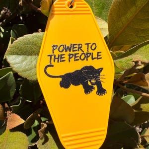 Puede incluir: Llavero amarillo estilo motel con la frase "POWER TO THE PEOPLE" en negro y un diseño de pantera negra. El llavero tiene una textura acanalada y un anillo metálico. El fondo es una vista borrosa de follaje verde.