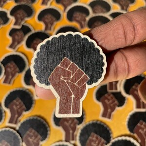 Può includere: Un ritaglio di legno di un pugno alzato con un'acconciatura afro nera. Il pugno è marrone e l'afro è nero. Il ritaglio è su uno sfondo giallo.