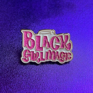 Könnte beinhalten: Eine goldfarbene Anstecknadel mit weißem Hintergrund und pinkfarbenem Glitzer-Schriftzug, der "Black Girl Magic" lautet.