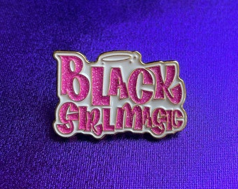 May include: A gold-colored pin with a white background and pink glitter lettering that reads "Black Girl Magic".