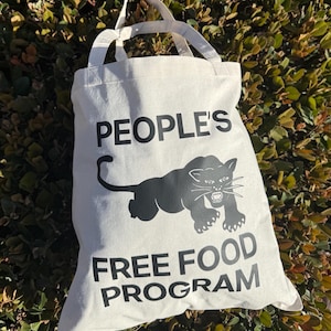May include: Off-white canvas tote bag with black text and a panther graphic. The text reads "PEOPLE'S FREE FOOD PROGRAM". The bag has two white handles and is set against a backdrop of green foliage.
