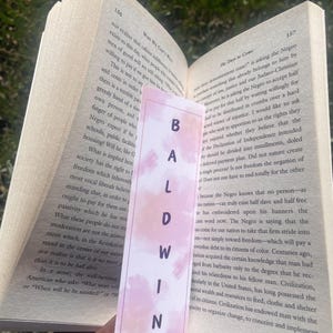 Może przedstawiać: Zbliżenie r&oacute;żowo-białego zakładki z napisem "BALDWIN" napisanym czarnymi literami. Zakładka znajduje się na stronie książki z widocznym tekstem "The Daye to Come" i "WHY WE CAN'T WAIT".
