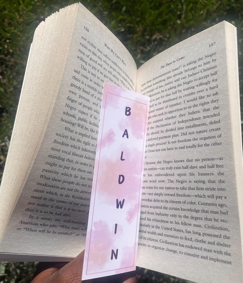 Może przedstawiać: Zbliżenie r&oacute;żowo-białego zakładki z napisem "BALDWIN" napisanym czarnymi literami. Zakładka znajduje się na stronie książki z widocznym tekstem "The Daye to Come" i "WHY WE CAN'T WAIT".