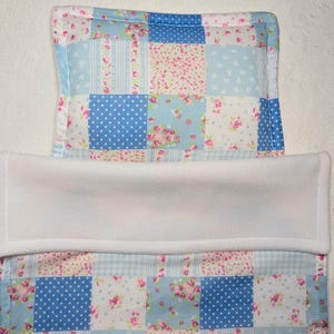 May include: A patchwork baby changing mat cover with a white fleece centre. The patchwork design features squares of blue, pink floral, and white polka dot fabrics. The edges are trimmed with a coordinating floral fabric.