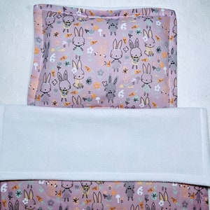 May include: A pink and white baby cot bedding set with a bunny rabbit pattern. The bedding includes a fitted sheet, a cot skirt, and a pillowcase. The pattern features bunnies, carrots, and flowers.