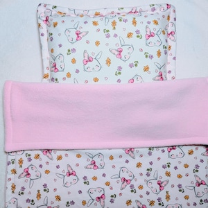 May include: A baby bedding set with a white fitted sheet and pillowcase, patterned with bunny faces wearing pink bows and flowers. A pink fleece blanket is folded over the top. The set is designed for a cot.