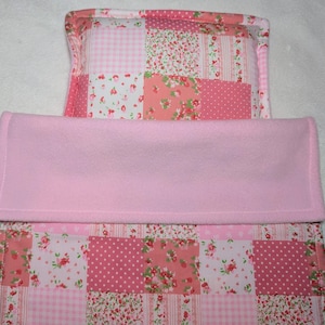 May include: A pink patchwork baby blanket and pillow set. The blanket has a soft pink fleece section and a patterned section with floral and polka dot designs in pink and white. The pillow has a similar patchwork design, creating a coordinated nursery set.