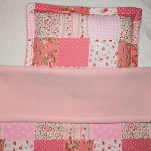 May include: A baby bedding set with a patchwork pillow and a blanket. The pillow features floral and polka dot patterns in pink and white. The blanket is a solid light pink colour, with a soft, plush texture.