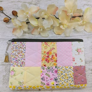 May include: A patchwork zipped pouch featuring floral and polka dot patterns in pink, yellow, and white. The pouch has a dark green zip and a tassel charm. The design is quilted with a diamond pattern.