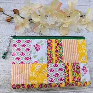 May include: A quilted patchwork pouch with a green zip and tassel. The pouch features a variety of floral and geometric patterns in pink, yellow, and white. The pouch is approximately 20 cm wide and 15 cm tall.