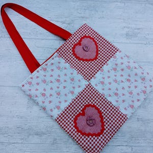 May include: A handmade tote bag with a red handle and a patchwork design. The bag features red and white gingham squares, floral print squares, and two heart-shaped appliqués with button details. The bag is made of fabric.