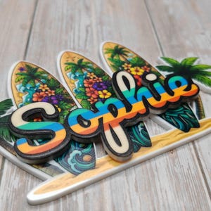 May include: A decorative sign featuring the name Sophie in a rainbow-striped font layered over three surfboard shapes. The surfboards are decorated with tropical palm trees, colourful hibiscus flowers, and ocean waves in shades of blue and green.