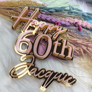 Op de afbeelding: Een gouden en roze acryl taarttopper met de woorden "Happy 60th" en de naam "Jacque". De topper heeft een gouden omtrek en een roze binnenkant, perfect voor een mijlpaal verjaardagsviering.