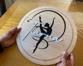 May include: A round wooden sign with a black silhouette of a gymnast holding a ribbon. The name "Tiana" is written in white 3D letters on the sign.