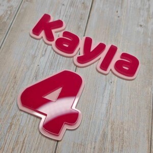May include: A layered acrylic sign featuring the name Kayla in a bold, dark pink font with a light pink backing, accompanied by a separate layered acrylic number 4 in the same dark pink and light pink color scheme with a glossy finish.