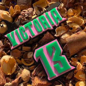 May include: Two pink and green name and number patches. One patch reads "VICTORIA" in green, with a black outline. The other patch has the number "13" in green, with a black outline. The patches are on a bed of dried botanicals.