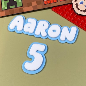 May include: A blue and white name banner with the name "AARON" and the number "5" in a playful, cartoon style. The banner is on a green background.