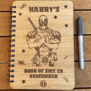 May include: Wooden spiral-bound notebook with a laser-etched Deadpool design. The notebook cover features the text "HARRY'S BOOK OF SHIT TO REMEMBER" and a Deadpool logo. A gray Sharpie pen is next to the notebook.