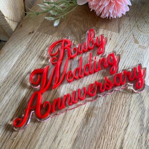Op de afbeelding: Een heldere acryl taarttopper met de woorden "Ruby Wedding Anniversary" in rood script. De topper is ontworpen voor een huwelijksverjaardag. De tekst is gelaagd over een heldere basis, waardoor een 3D-effect ontstaat.