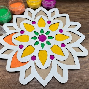 May include: A white wooden mandala with cutouts for sand art. The mandala has a center circle with a green leaf design and a purple dot. The surrounding sections are filled with yellow, orange, and purple sand.