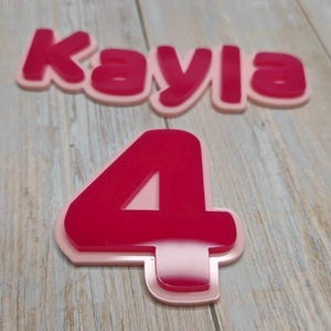 May include: A layered name sign reading Kayla in a bold, rounded font and a separate number four, both featuring a glossy dark pink top layer mounted onto a solid light pink base layer, creating a dimensional effect with clean, smooth edges.