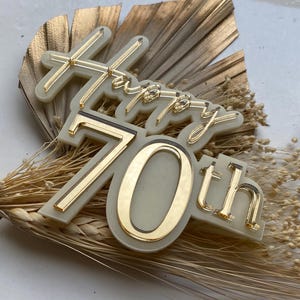 Op de afbeelding: Een witte acryl taarttopper met gouden letters die "Happy 70th" zeggen.