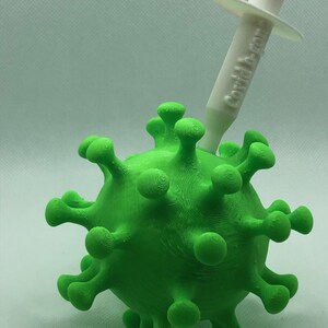 May include: A green 3D printed model of a coronavirus with a white syringe on top. The syringe has the text "COVID-19 VACCINE" printed on the side.