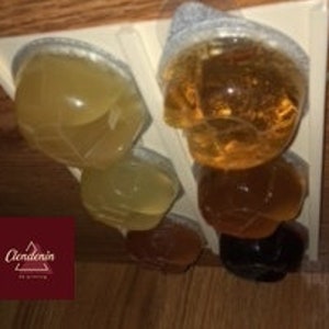 May include: Four clear plastic cups filled with different colored liquids. The cups are arranged in two rows of two. The top row has a light yellow liquid and a light brown liquid. The bottom row has a darker yellow liquid and a dark brown liquid.