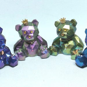 Op de afbeelding: Vier geometrische teddybeerfiguurtjes met iriserende afwerkingen. Elke beer draagt een gouden kroon en heeft een gouden ketting om zijn nek. De beren zijn in tinten paars, groen en blauw.