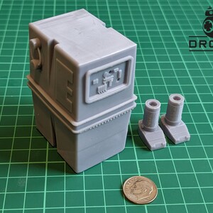 以下が含まれることがあります： スターウォーズのR2-D2ユニットのグレーのプラスチックモデルで、グレーのプラスチック製の脚が2本別々に付いています。モデルは緑色のカッティングマットの上にあり、アメリカの1セント硬貨がスケールとして置かれています。