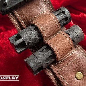 May include: A close-up shot of a brown leather belt with two dark grey cylindrical objects attached. The objects are held in place by smaller brown leather straps. The belt has white stitching and a brass-coloured stud.
