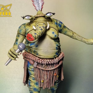 Può includere: Una statuetta dipinta di verde e blu di una creatura con un lungo muso, occhi grandi e un copricapo piumato. La creatura tiene un microfono e indossa una gonna di corda marrone.