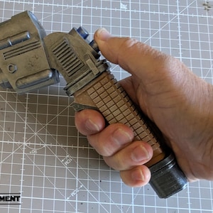 May include: A metallic grey and brown prop, possibly a futuristic device or tool, held in a hand. The device has a textured handle and various mechanical details, suggesting a sci-fi or industrial theme. The logo "EQUIPMENT" is visible in the bottom left corner.