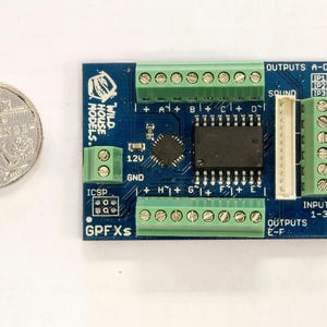 May include: A blue circuit board with green connectors, a black chip, and the text "GPFXs". A coin is next to the board for size comparison. The board has the text "WILD HOUSE MODELS" and "OUTPUTS A-D".