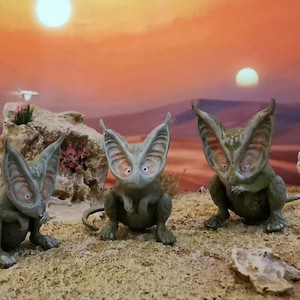 May include: Three whimsical, green-toned creature figurines with large ears and wide eyes are posed on a sandy surface. The background features a sunset with two suns and a desert landscape, creating a fantasy scene.