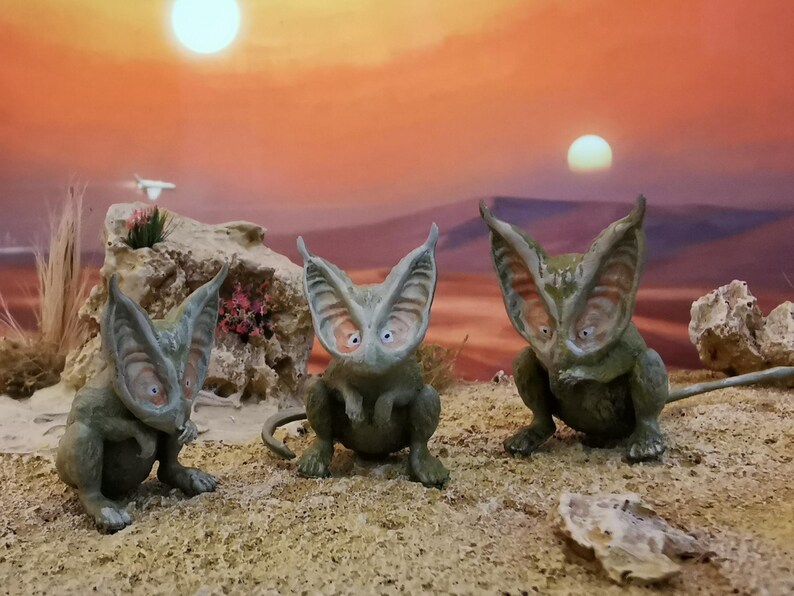May include: Three whimsical, green-toned creature figurines with large ears and wide eyes are posed on a sandy surface. The background features a sunset with two suns and a desert landscape, creating a fantasy scene.