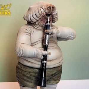 Può includere: Una statuetta bianca e grigia di un personaggio di Star Wars che suona un clarinetto nero. Il personaggio ha una testa grande con un viso bianco e rugoso e un naso grande e rotondo. Il personaggio indossa una camicia bianca e pantaloni marroni.