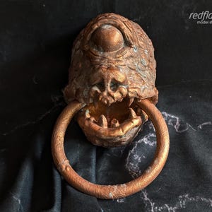 Peut inclure: Heurtoir de porte en bronze en forme de monstre cyclope avec un grand œil et une bouche ouverte. Le heurtoir a une poignée en forme d'anneau et est posé sur un fond sombre et texturé. Le texte "redflare model studios" est dans le coin supérieur droit.