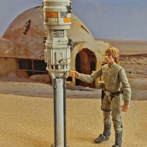 May include: A white and orange Star Wars action figure of Luke Skywalker standing in a desert setting next to a white and grey droid. The droid is on a tall, white pole.