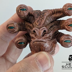 Pode incluir: Close-up de uma criatura de vários olhos pintada à mão, de cor castanho e bege, com olhos turquesa. A criatura tem uma superfície texturizada e é esculpida em um estilo realista.