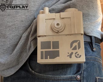 May include: A beige rectangular prop with a series of geometric shapes and symbols, attached to a person's belt. The prop has a cylindrical design on top. The word "COSPLAY" is visible in the upper left corner.