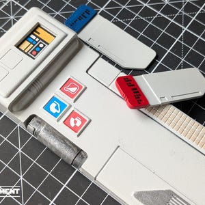 Op de afbeelding: Een close-up van een wit en grijs apparaat met een scherm en gekleurde knoppen. Het apparaat heeft een rechthoekige vorm met een scherm dat een afbeelding weergeeft. Er zijn rode, blauwe en witte accenten. Het woord "EQUIPMENT" is zichtbaar.