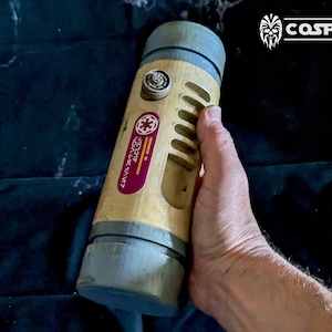 Op de afbeelding: Een grijs en beige cilindrisch rekwisiet met een rood en oranje label met de tekst "STAR WARS 4L02H". Het rekwisiet heeft een kleine knop aan de bovenkant en een reeks sleuven aan de zijkant.