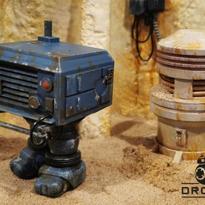 Op de afbeelding: Een blauw en grijs robot speelgoed met een roestige bruine metalen pijp op de achtergrond. De robot heeft een vierkant lichaam met een rooster aan de voorkant en een kleine antenne bovenop. De pijp heeft een roestige bruine metalen bovenkant en een witte basis.