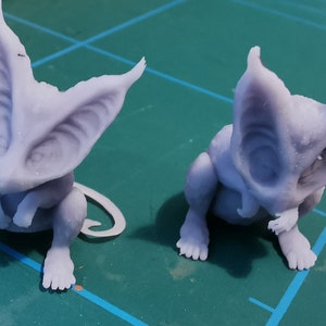 May include: Two small, light purple, 3D printed figurines of a creature with large ears and a curled tail. The creature is sitting with its paws tucked under its chin.