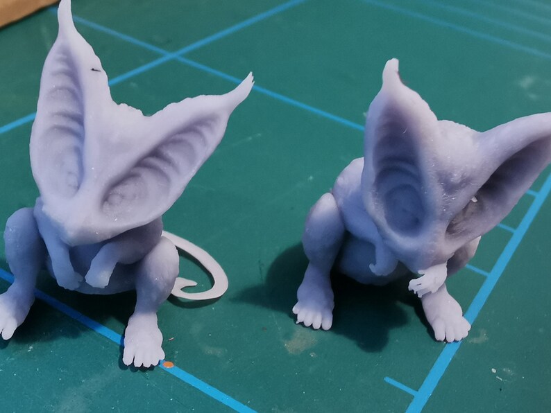 May include: Two small, light purple, 3D printed figurines of a creature with large ears and a curled tail. The creature is sitting with its paws tucked under its chin.